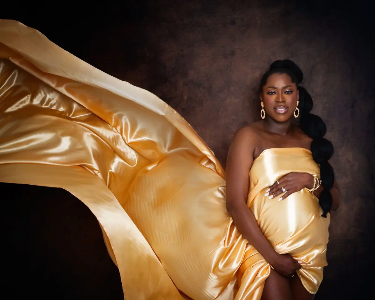 a pregnant woman stands against a deep brown background with a golden fabric flying out behind her