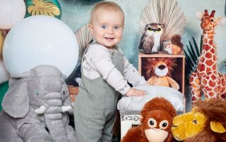 a little boy stands in front of a jungle themed backdrop with some jungle stuffed toys.