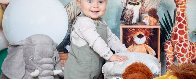 a little boy stands in front of a jungle themed backdrop with some jungle stuffed toys.