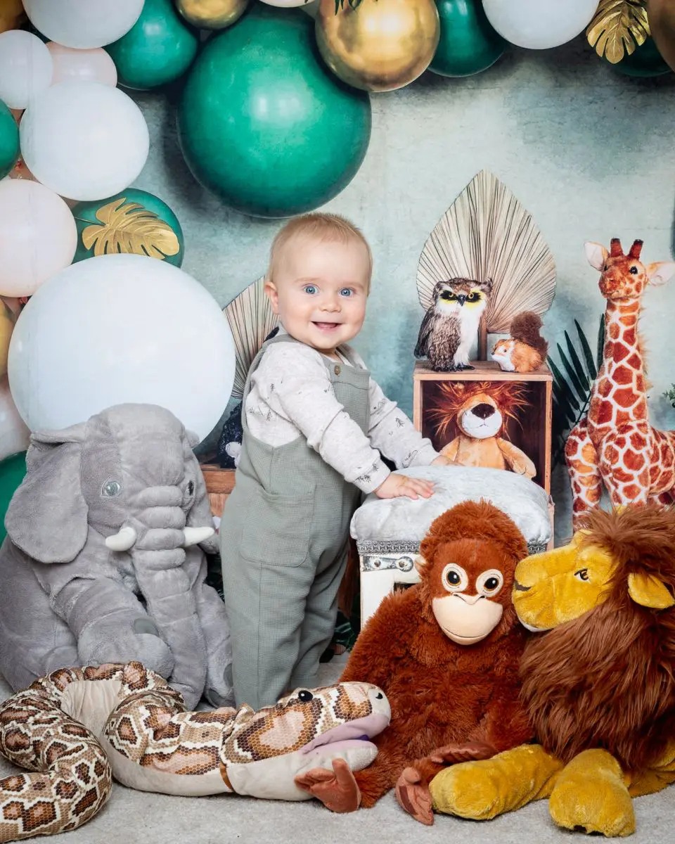 a little boy stands in front of a jungle themed backdrop with some jungle stuffed toys.