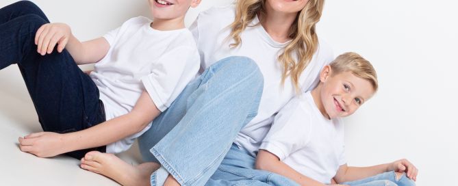 A mum in jeans and white tshirt sits on the ground between her two sons