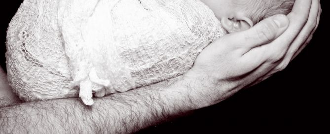 a swaddled sleeping newborn photographed in dad's arms against a black background in a black and white image