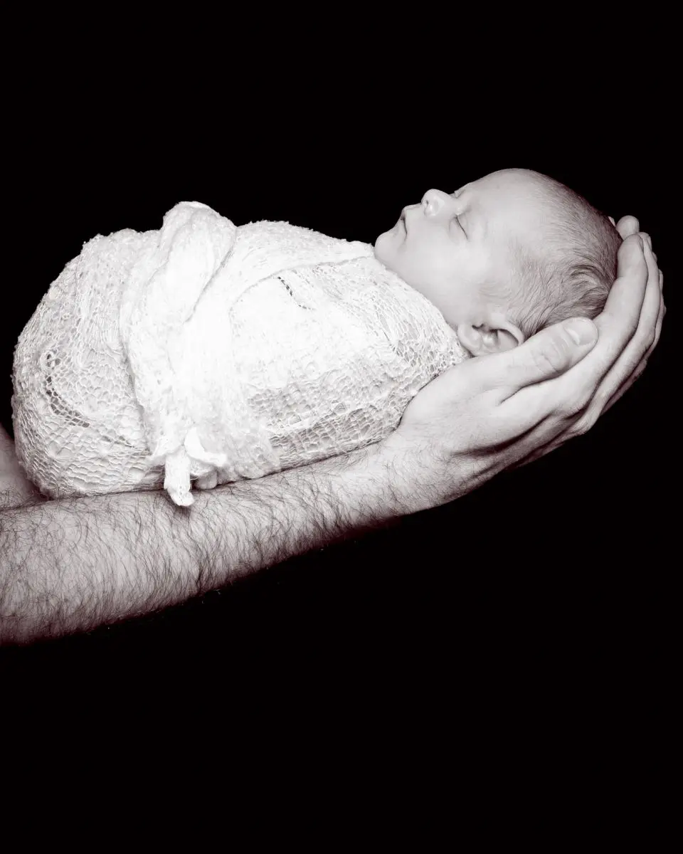 a swaddled sleeping newborn photographed in dad's arms against a black background in a black and white image