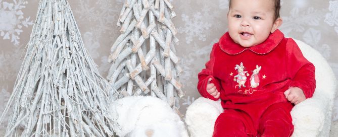 A smiling baby in a red romper poses with Christmas props at a photo shoot