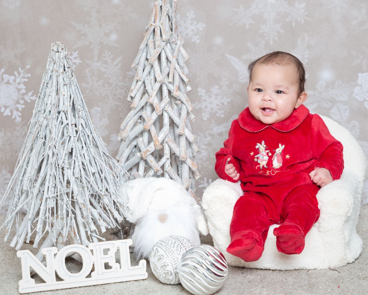 A smiling baby in a red romper poses with Christmas props at a photo shoot