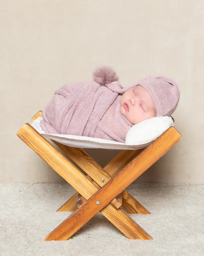 a newborn girl sleeps in a wooden hammock in a dusty rose coloured wrap with matching hat.