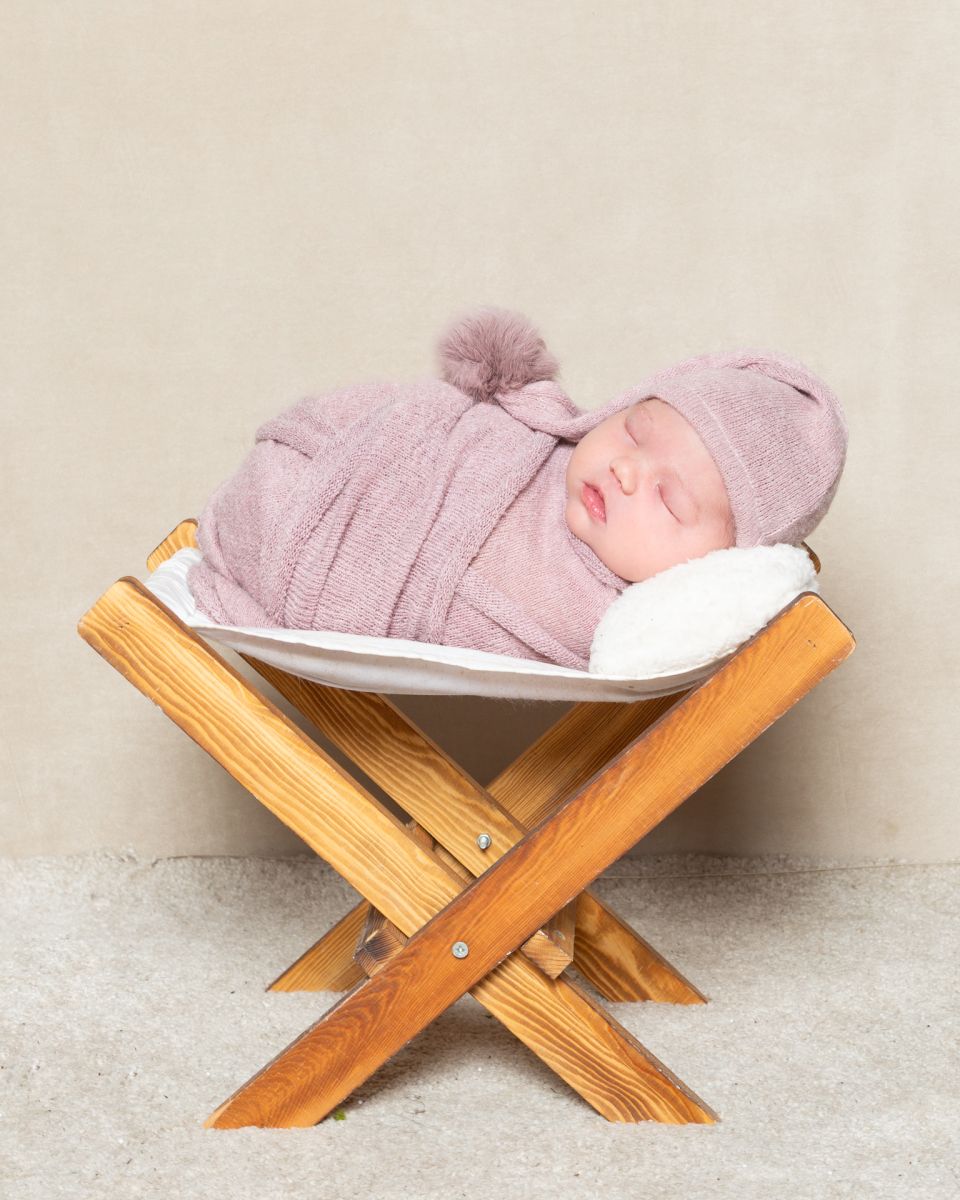 a newborn girl sleeps in a wooden hammock in a dusty rose coloured wrap with matching hat.