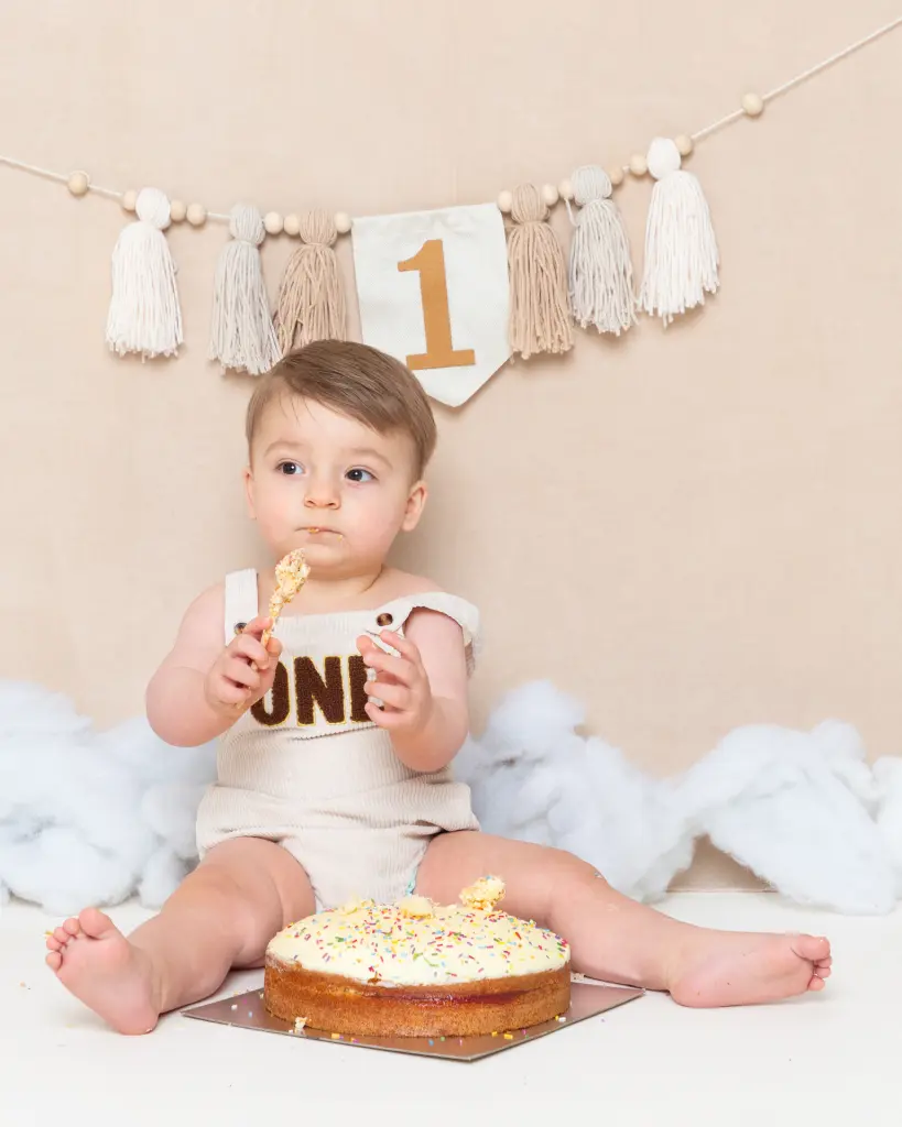 a little boy eating cake dressed in a cream outfit against a neutral background at a cake smash photo shoot