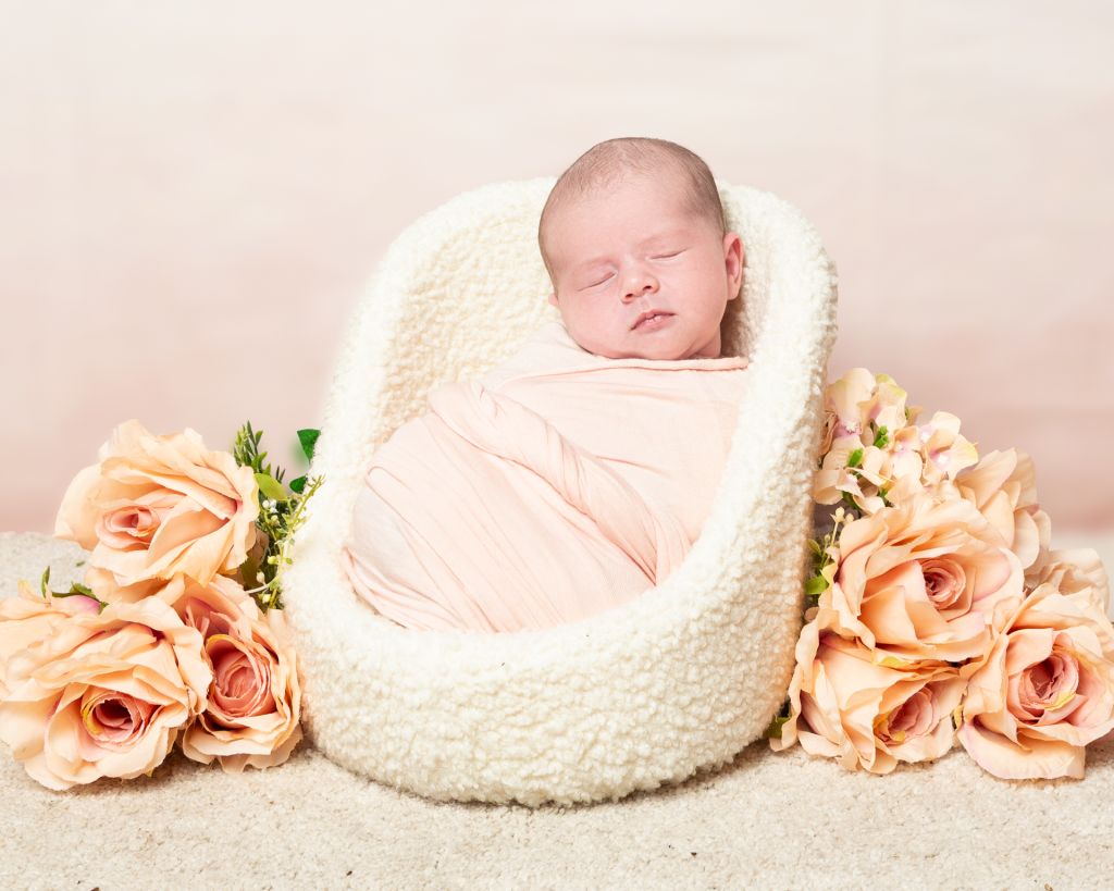 a little girl swaddled in peach sleeps in a chair next to peach flowers