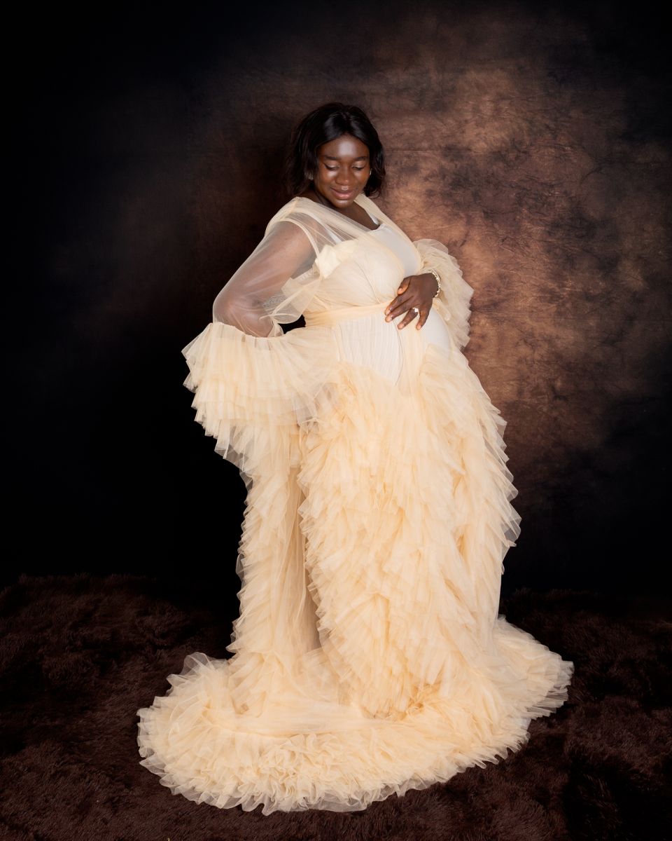a woman poses against a brown fine art backdrop in a cream tulle robe