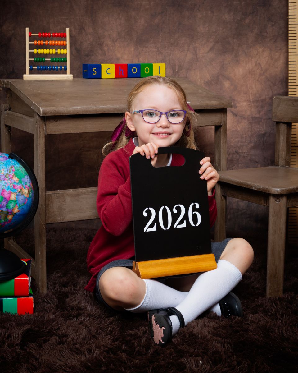 a little girl in a red school jumper holds a sign at photo shoot