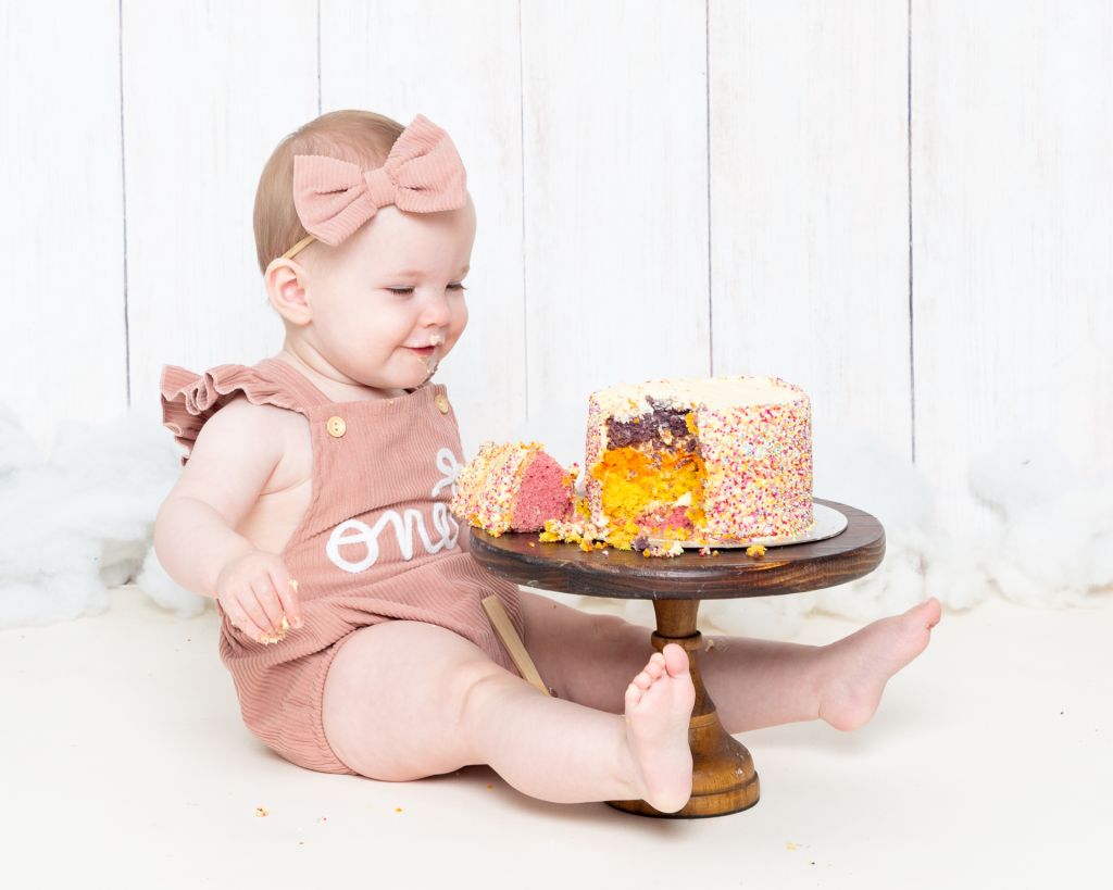 a little girl in a pink romper with a rainbow cake