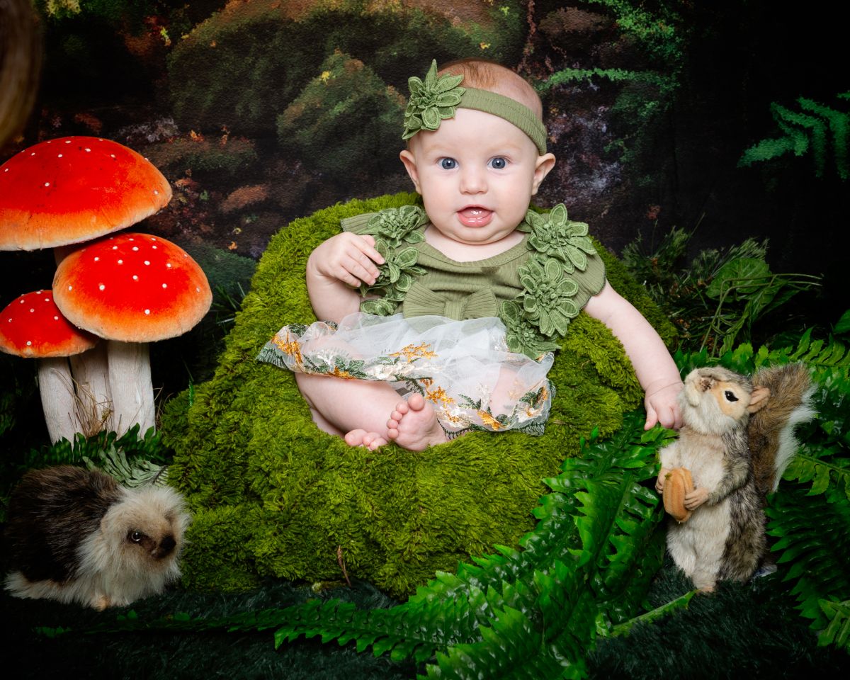 a baby girl sits in a moss covered chair in a woodland themed photo shoot